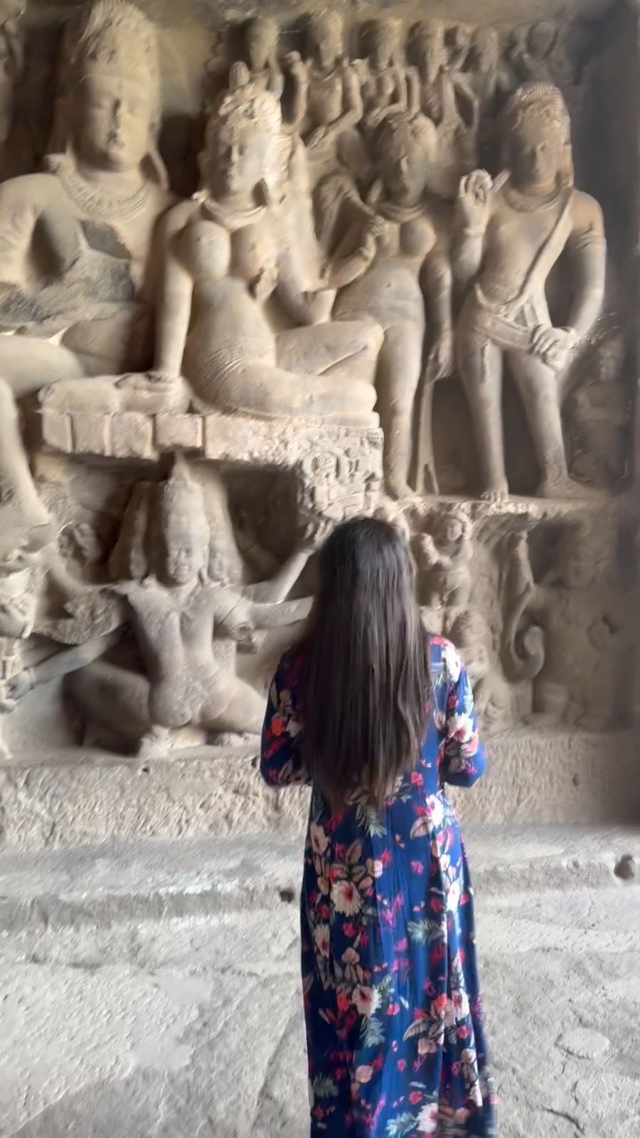 Standing before a carved wall of deities in an Ellora cave.